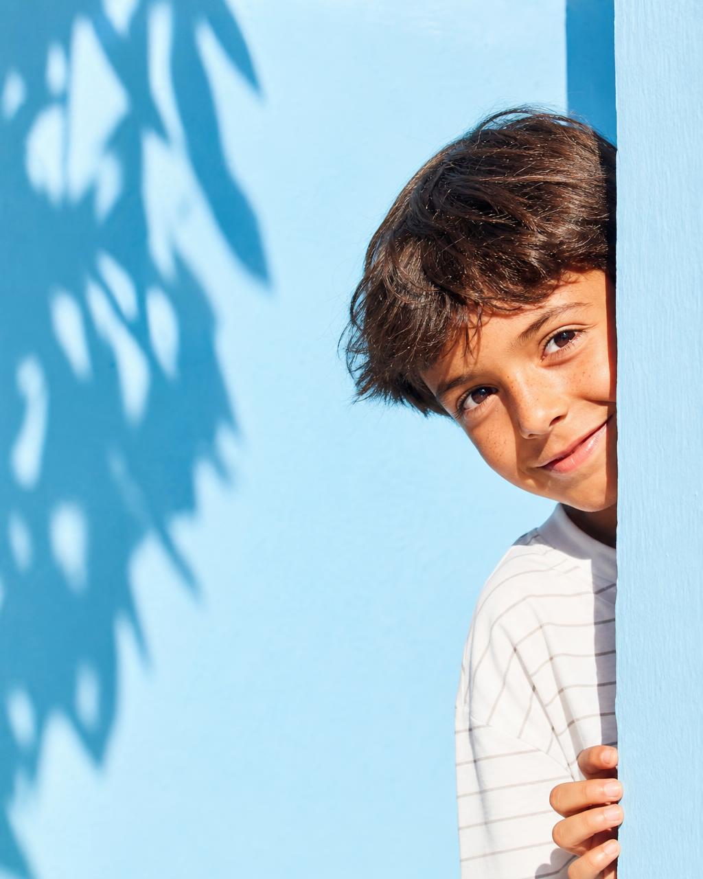Niño asomando por el borde del encuadre sobre fondo azul claro con sombra de hojas
